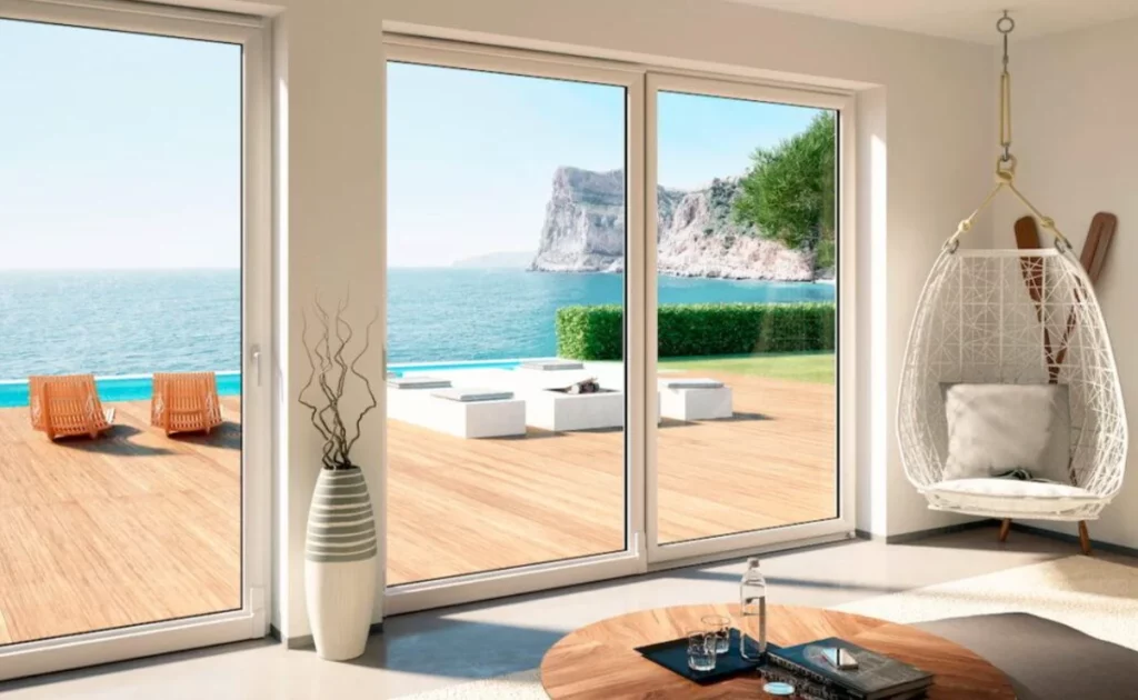 Interno luminoso con grandi finestre scorrevoli che affacciano su un paesaggio marino con scogliere e una piscina.