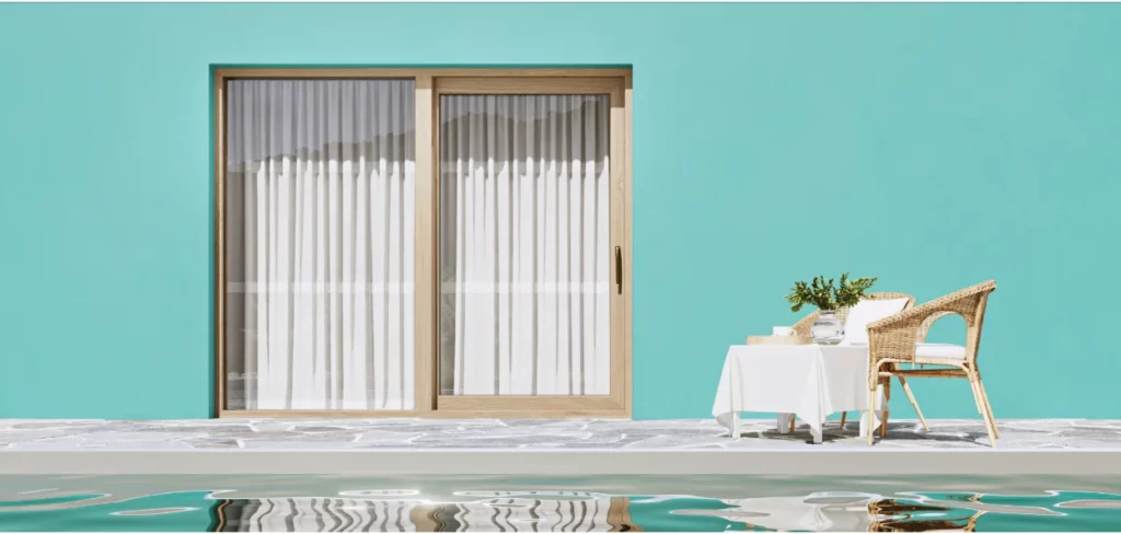 Porta scorrevole in legno con tende bianche, su parete color acquamarina. Tavolino e sedie in vimini vicino a piscina riflettente.