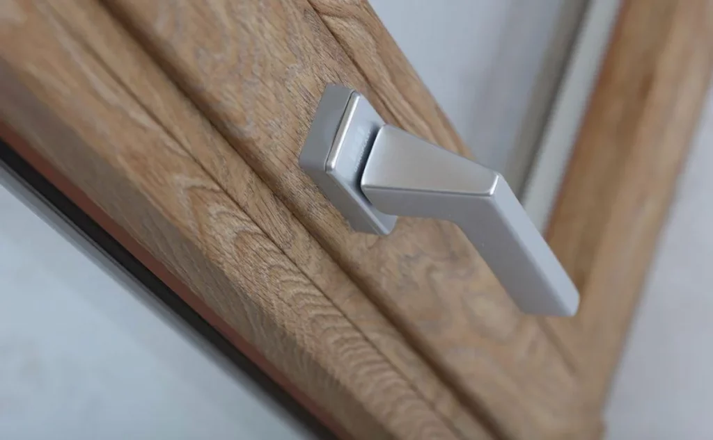 Dettaglio maniglia in metallo satinato su telaio finestra in legno chiaro con venature evidenti.